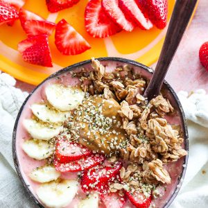 Easy Strawberry Banana smoothie bowl in coconut bowl with fresh strawberries, bananas, granola, peanut butter, and hemp hearts with tan napkin.