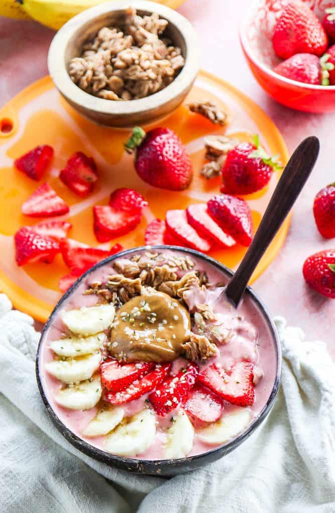 Strawberry Banana Smoothie bowl with fresh fruit, homemade granola on a pink backdrop with tan napkin 