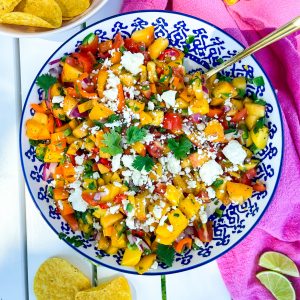 Sweet and spicy peach mango salsa in a blue and white bowl topped with fresh cilantro and feta cheese and served with tortilla chips.