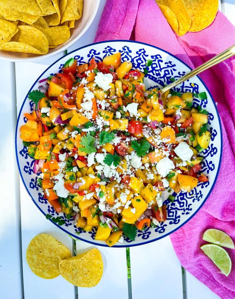 Sweet and spicy peach mango salsa in a blue and white bowl topped with fresh cilantro and feta cheese and served with tortilla chips. 