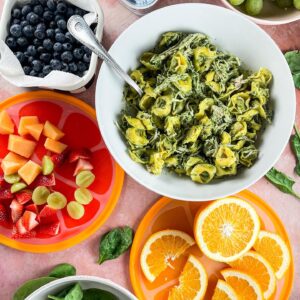 chicken and pesto tortellini salad in a white bowl with fresh fruit and salad